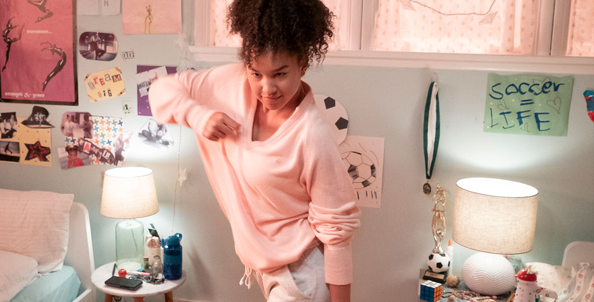 A teenage girl wearing bright pink clothing dances in her bedroom which is also colorful and adorned with inspirational words.
