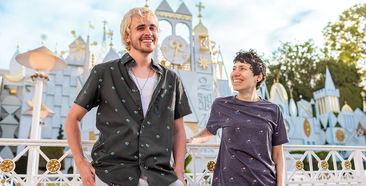Two people stand in front of it’s a small world, smiling and wearing Disney Pride Collection shirts. The person on the left wears a shirt with rainbow insignias for both the Rebel Alliance and Galactic Empire, while the person on the right wears a shirt with rainbow versions of the Pixar Lamp.