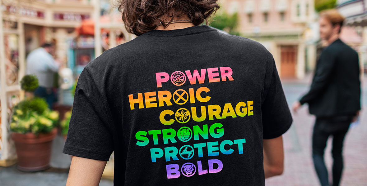 A person faces away from the camera, wearing a black T-shirt with rainbow text that says, “POWER, HEROIC, COURAGE, STRONG, PROTECT, BOLD.”