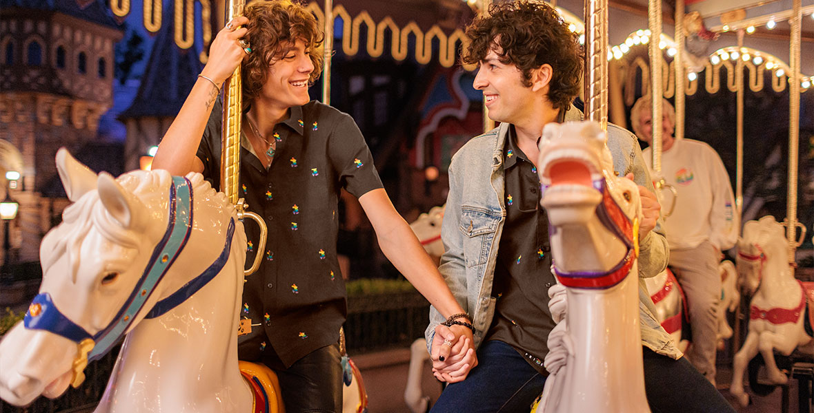 A couple holds hands on King Arthur’s Carousel, wearing matching polo shirts decorated with rainbow silhouettes of Mickey Mouse.