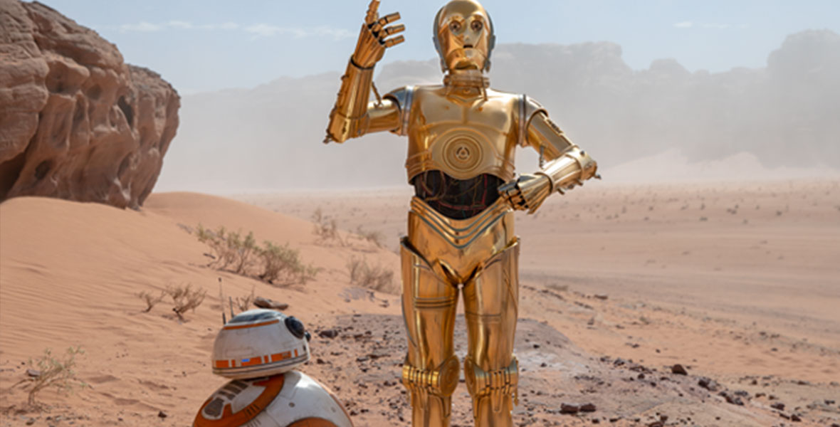 Standing in a desert, C-3PO (Anthony Daniels) raises his gold arm in exasperation while BB-8 looks on.