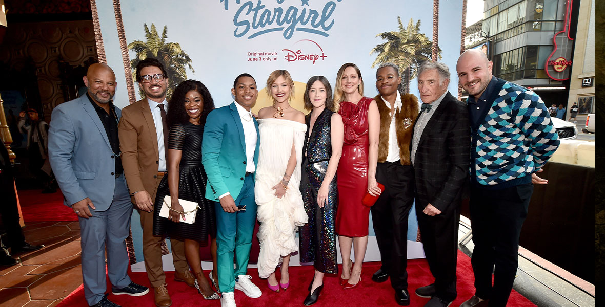 The cast and crew of Disney+’s Hollywood Stargirl pose together on the red carpet at the El Capitan Theatre in front of a large poster for the film.