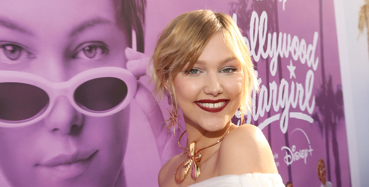 Grace VanderWaal smiles on the red carpet in front of a large poster of herself in Hollywood Stargirl. She has her short blond hair pulled back and is wearing a white off-the-shoulder dress and large gold accessories.
