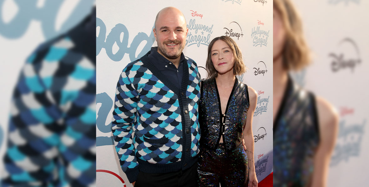 Director and co-writer Julia Hart and writer Jordan Horowitz pose together on the red carpet. Horowitz is wearing a scale-patterned button-up sweater and Hart is wearing a sparkly, holographic matching pant and vest set.