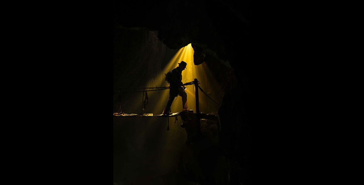 Indiana Jones stands silhouetted in profile, with golden light shinning down on him.