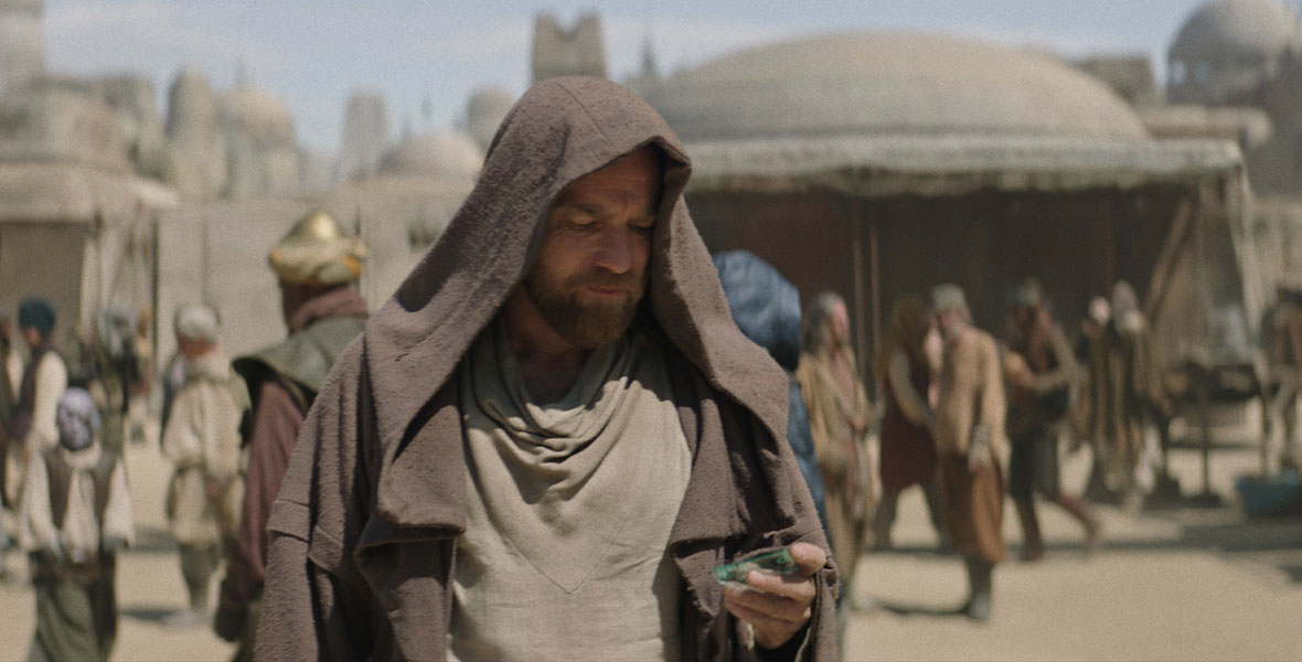 Ewan McGregor, as Obi-Wan Kenobi, stands in a Tatooine marketplace, covered by his cloak. He looks down at something in his hand.