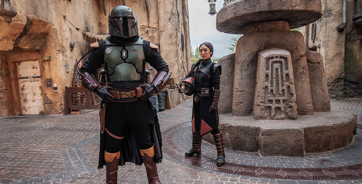 Boba Fett, with his helmet on, and Fennec Shand, holding her helmet in her arm, stand inside the Star Wars: Galaxy’s Edge marketplace.