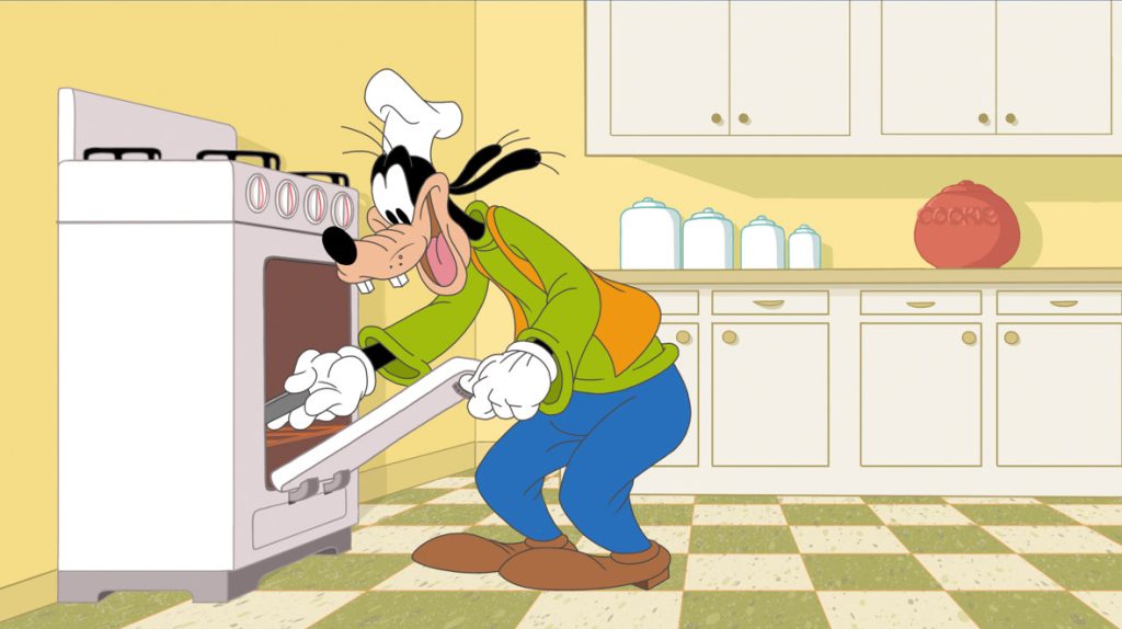 Goofy is wearing a chef’s hat, a green shirt, orange vest, and blue pants, and he’s putting something into the oven in the kitchen, in a still from the “Learning to Cook” episode of How to Stay at Home.