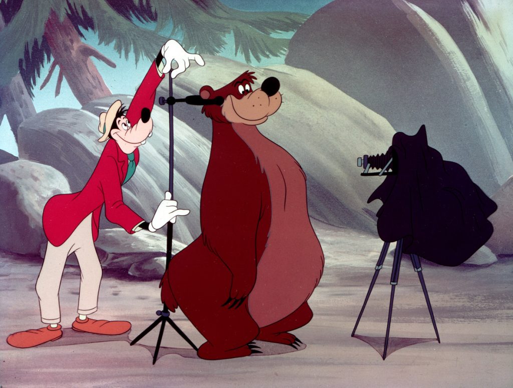 Goofy, wearing a red blazer and tan pants, is measuring a bear for a photo in a still from the animated short Hold That Pose. There is camera equipment nearby.