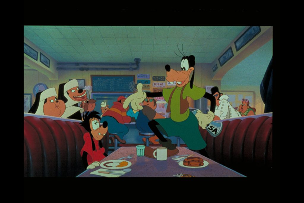 Max and Goofy are inside a crowded diner in a still from A Goofy Movie. There are two nuns, as well as someone dressed like Elvis. There are several plates of food on the table. Max is wearing a red shirt; Goofy is wearing a green shirt and shorts and a brown vest, and is holding a map of the United States.