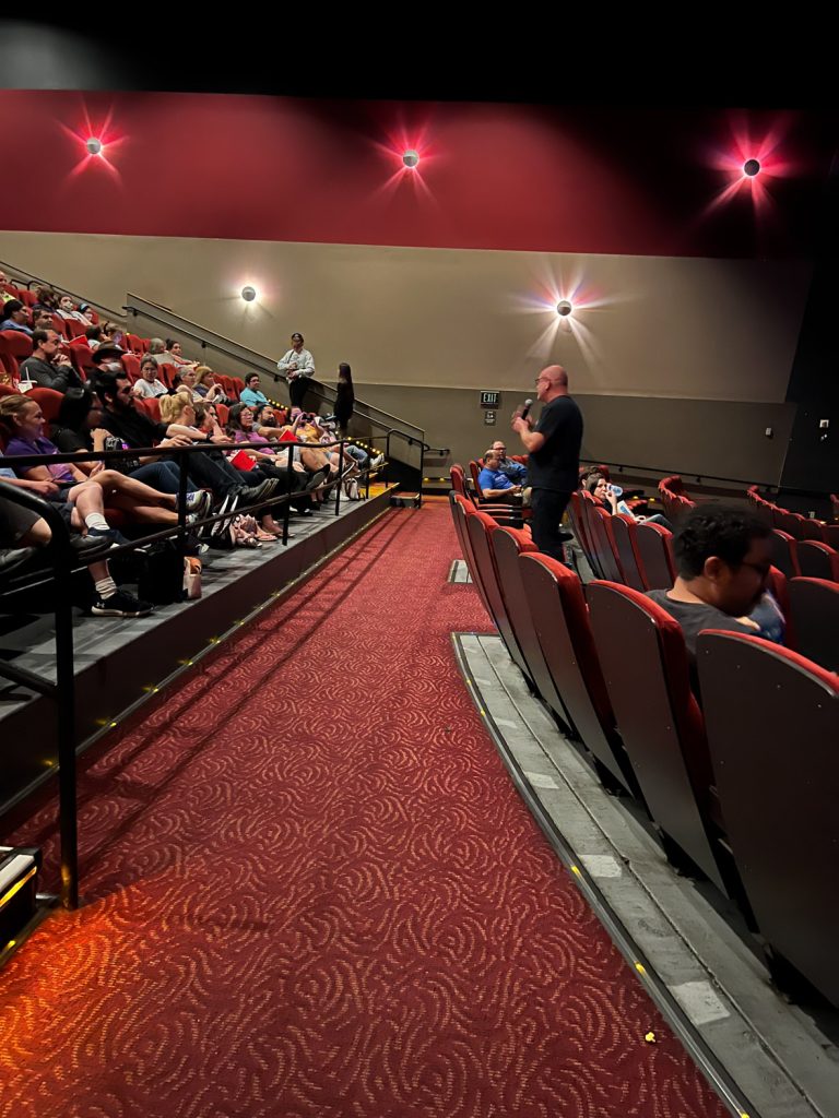 Head of D23 Michael Vargo stands among red movie theater seats holding a microphone, addressing the D23 Members sitting in front of him, before the screening beings.