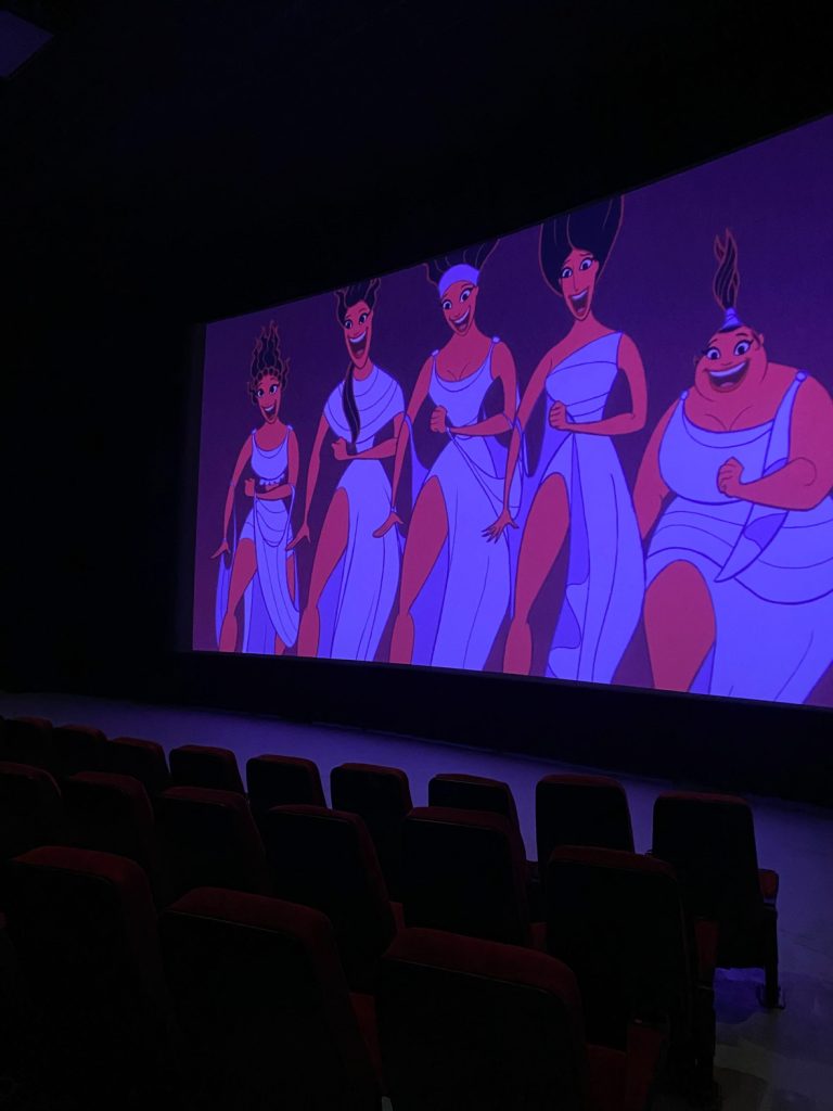 A still from the film Hercules on the movie theater screen, depicting the five Muses mid-song against a purple background.