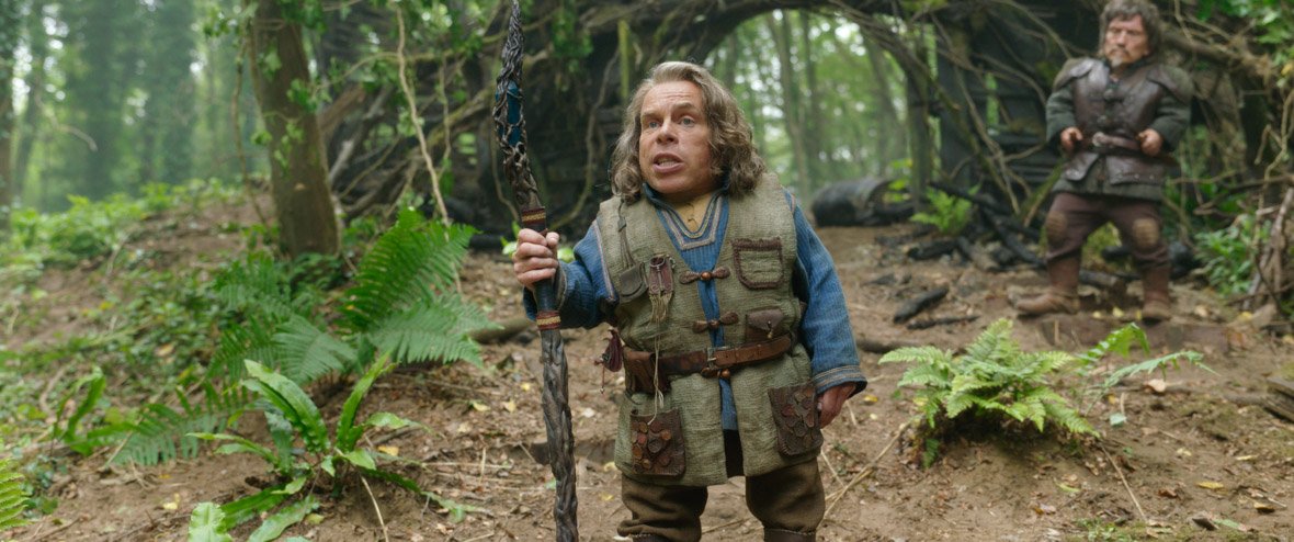 Warrick Davis as Willow Ufgood, standing in a forest and holding an intricately-carved wooden staff. He is mid-speech, talking to someone offscreen