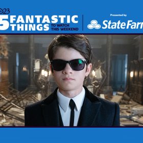 Artemis Fowl (Ferdia Shaw) dons a suit and tie with matching black sunglasses as he stands in an entryway that is destroyed with shattered glass on the floor and damaged furniture sprawled around the room in Disney’s Artemis Fowl.