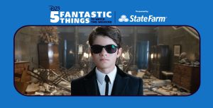 Artemis Fowl (Ferdia Shaw) dons a suit and tie with matching black sunglasses as he stands in an entryway that is destroyed with shattered glass on the floor and damaged furniture sprawled around the room in Disney’s Artemis Fowl.
