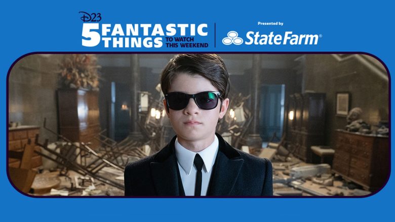 Artemis Fowl (Ferdia Shaw) dons a suit and tie with matching black sunglasses as he stands in an entryway that is destroyed with shattered glass on the floor and damaged furniture sprawled around the room in Disney’s Artemis Fowl.
