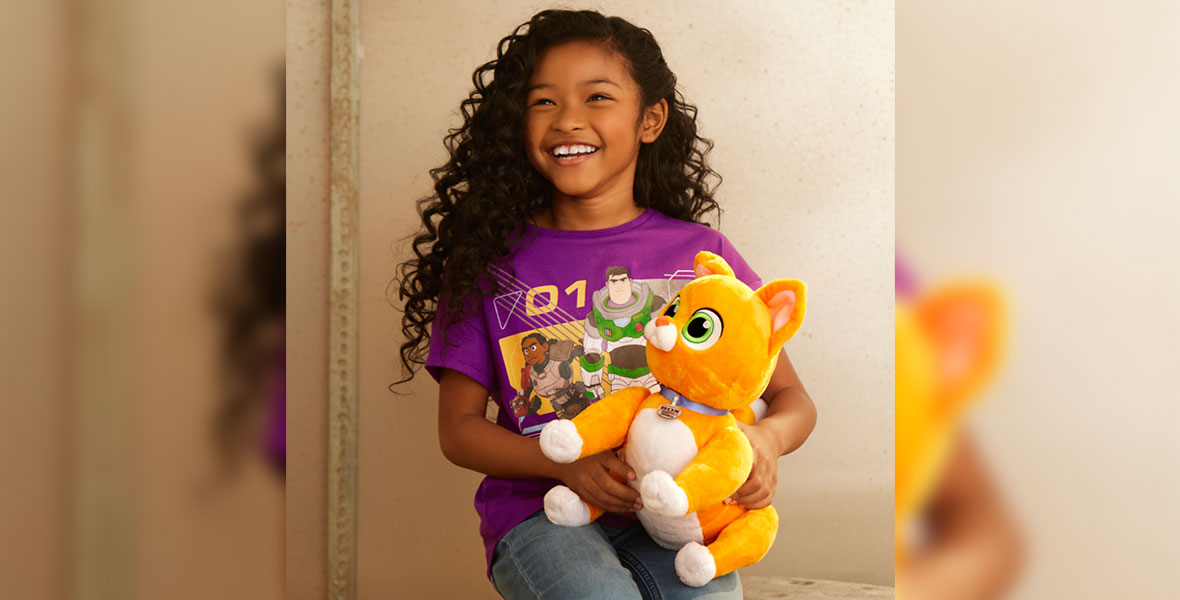 A young girl in a purple Lightyear shirt smiles as she holds a plush version of the character Sox, a robot cat, from Lightyear.