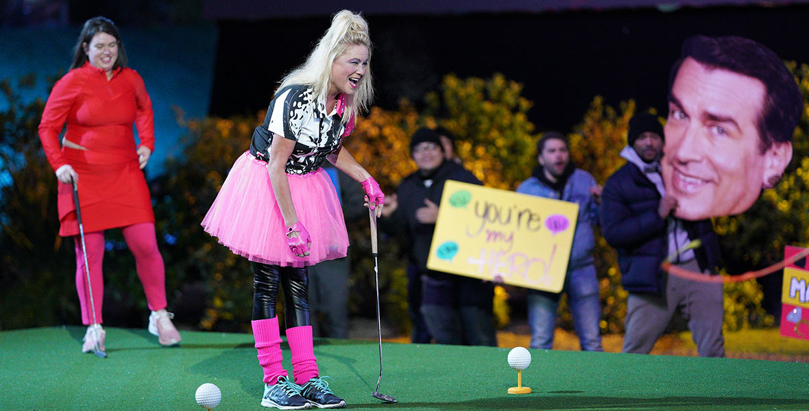 Two female contestants on the mini golf competition series Holey Moley stand on the putting green as they hold their golf clubs and look at the tees. The contestant on the left is wearing a red top and matching skirt with hot pink tights. The other contestant is wearing a black graphic tee with a hot pink tutu, black leggings, and hot pink legwarmers. Behind them stands the audience including a large cutout of Rob Riggle’s face.