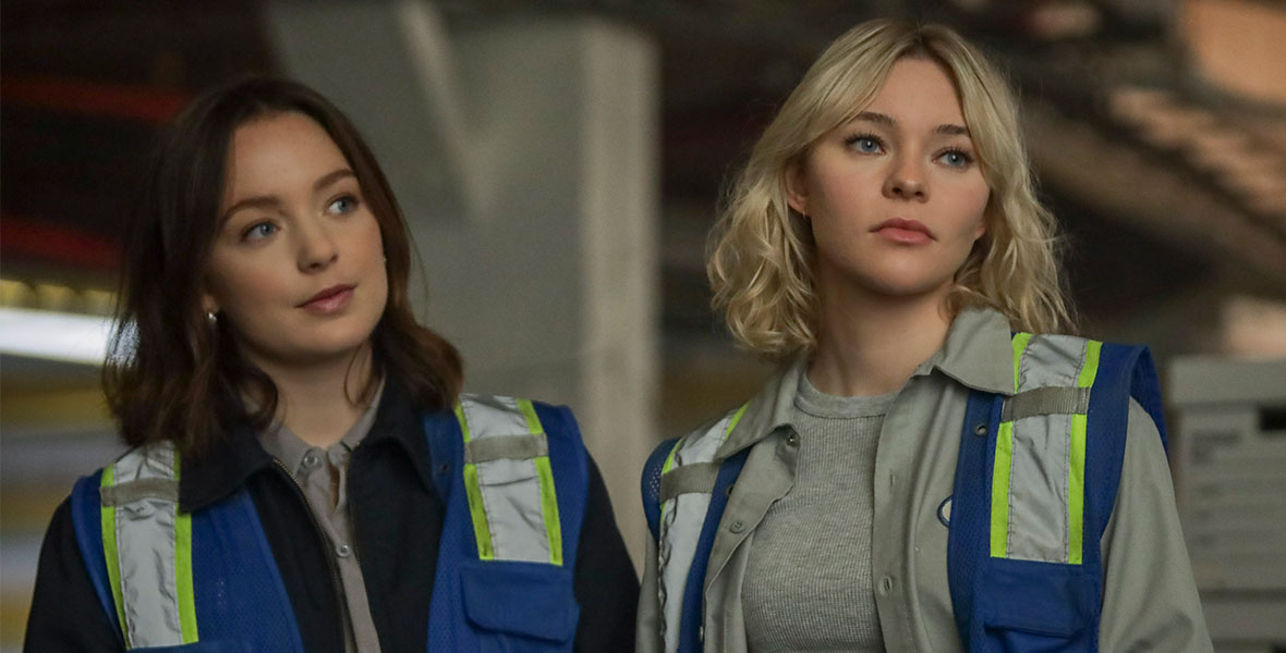 Scylla (Amalia Holm) and Raelle (Taylor Hickson) stand shoulder to shoulder wearing grey and black neutral clothing, and blue vests with yellow reflective accents, in the first episode of Motherland: Fort Salem season three. 