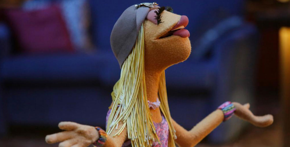 Janice stands with her arms outstretched, looking up. She wears a floral tank top, rainbow bracelet, and purple hat.