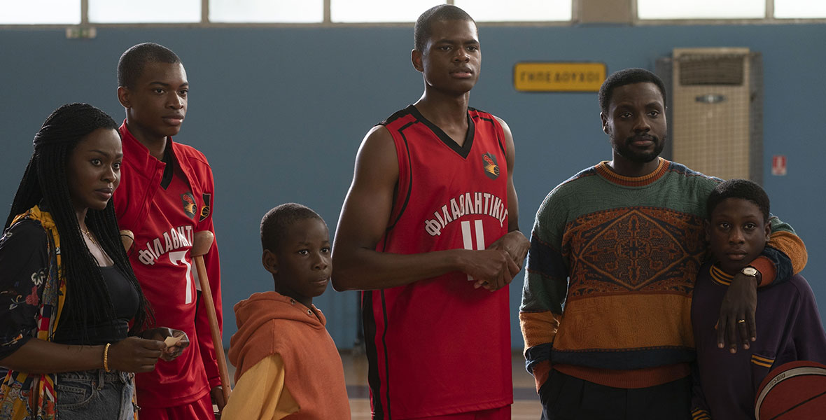 The Antetokounmpo family as depicted in Disney’s Rise stand huddled together in a brightly decorated gymnasium following Thanasis and Giannis’ basketball game. The brothers are wearing red jerseys with the team name emblazoned across the chest.