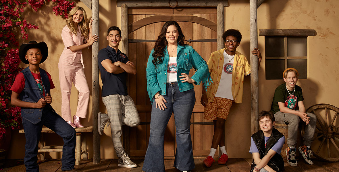 The cast of Disney Channel’s BUNK’D stands in front of a doorway to a dwelling; above the door is a wooden sign that reads “Kikiwaka Ranch.” Miranda May, who stars as Lou, stands in the middle of the group and is wearing a turquoise jacket with fringe and jeans. 