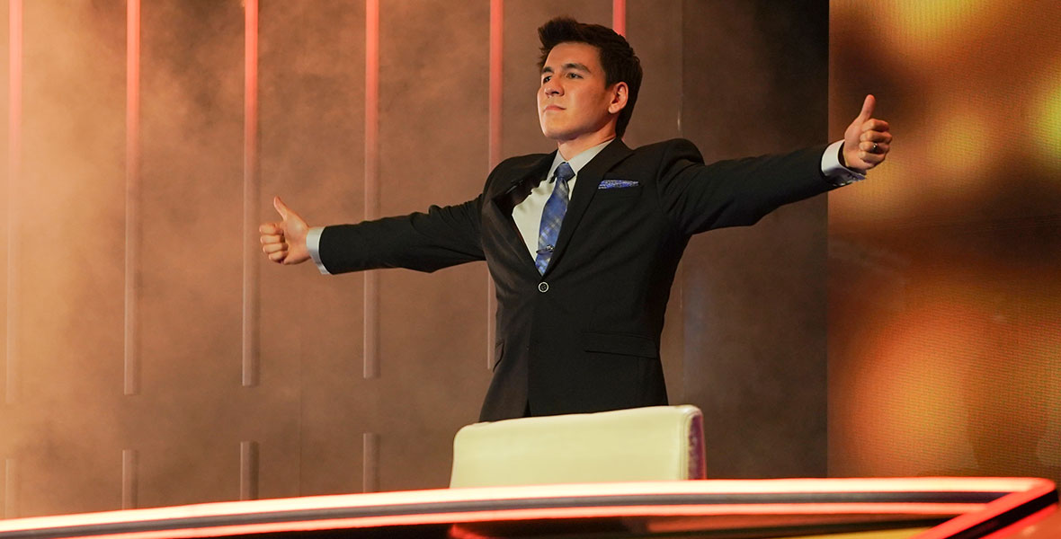 James “The High Roller” Holzhauer stands with his arms extended and thumbs up in front of large digital screens on the competition series The Chase.