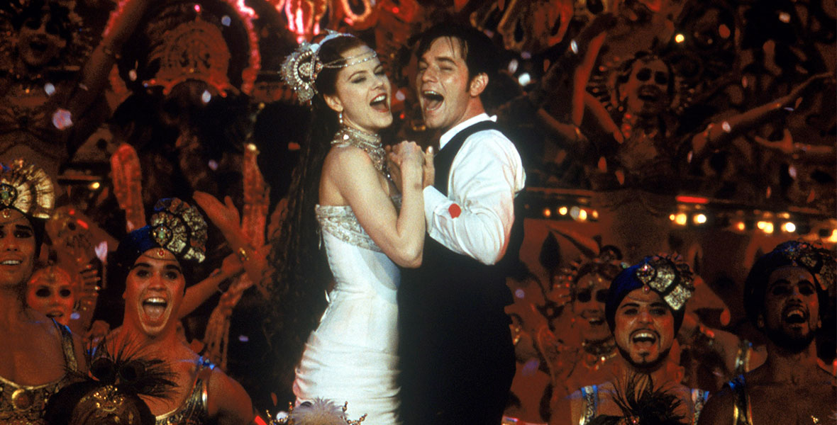 Satine (Nicole Kidman) in a white dress and sparkly headdress, is holding hands and standing side-by-side with Christian (Ewan McGregor), who’s wearing a white shirt with a black vest and pants. They’re singing, and surrounded by fancy-dressed dancers on all sides, in an image from Moulin Rouge!