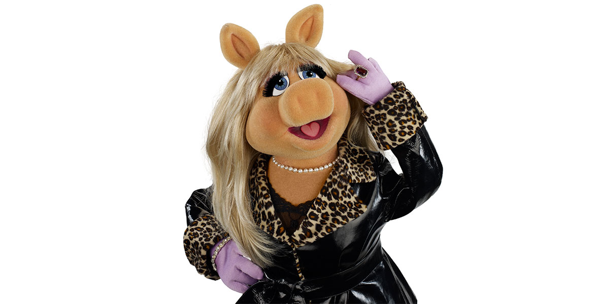 Miss Piggy strikes a pose with one hand on her hip and one hand against her head. She wears a black coat with leopard print accents, a pearl necklace, purple gloves, and a large ring.