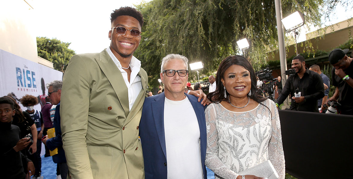 Producer Bernie Goldmann stands in between Giannis Antetokounmpo (left) and his mother Veronica Antetokounmpo (right). Giannis is wearing an olive green suit and sunglasses. Bernie has black square frame glasses and a blue blazer. Veronica has on a sparkly silver and white dress.