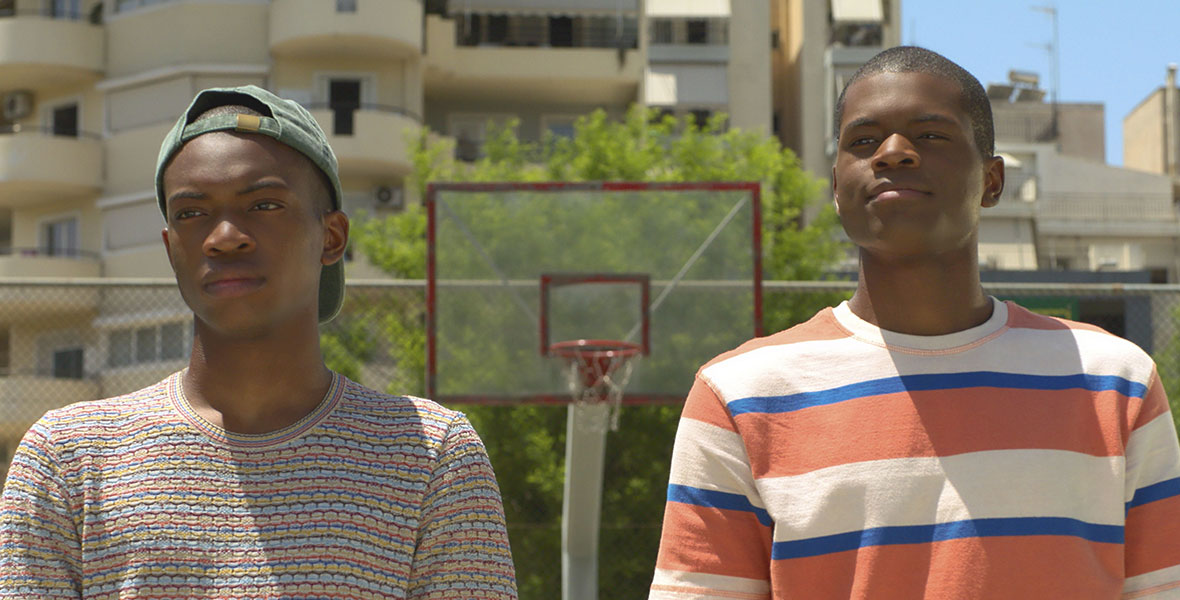 Thanasis (Ral Agada) and Giannis Antetokounmpo (Uche Agada) stand side by side while looking on at a group of boys playing basketball in park in Disney’s Rise.