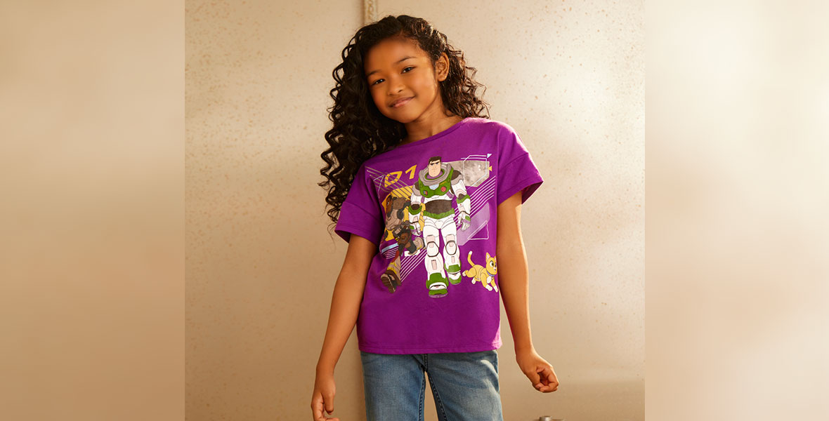 A young girl poses with a slight smile in a purple T-shirt featuring a 2D drawing of Izzy, Buzz Lightyear, and Sox.