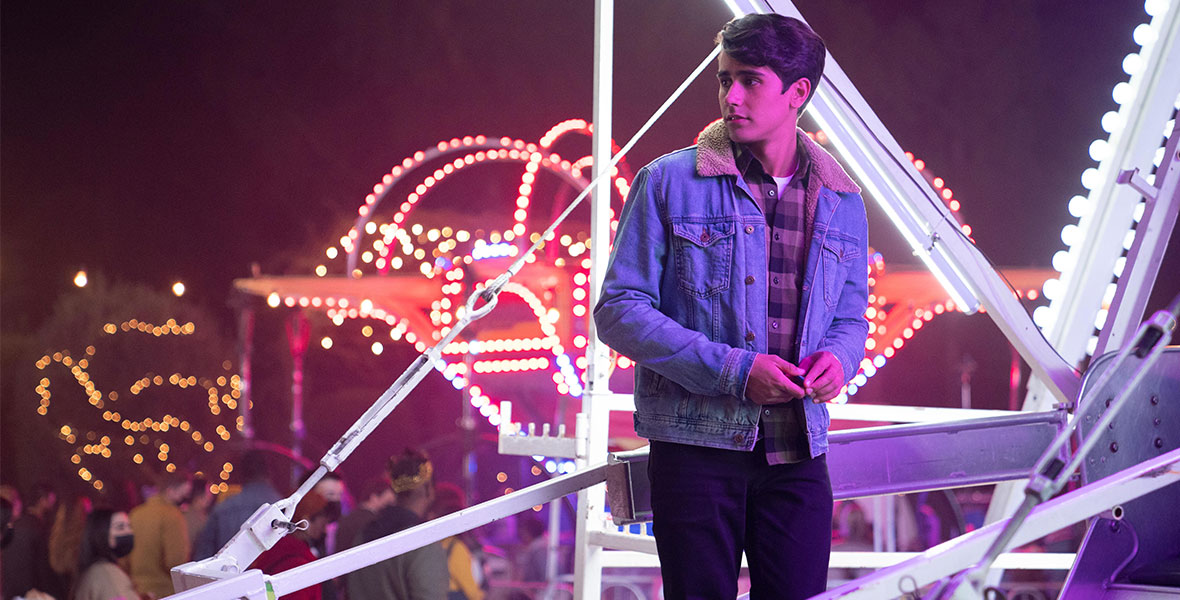 Victor (Michael Cimino) in Hulu Original series Love, Victor looks off at the crowd as he stands next to a Ferris wheel at the Winter Carnival.