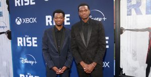 Ral Agada is wearing a blue and black patterned suit. Next to him is his brother, Uche Agada, wearing an all black paisley patterned suit. The pair stand with their hands clasped in front of them with a &ldquo;Rise x XBOX&rdquo; background behind them.