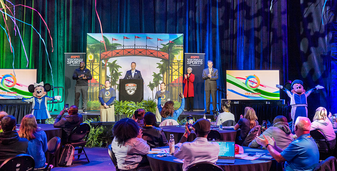 A stage with Mickey Mouse and Minnie Mouse standing on opposite sides with their hands in the air as Walt Disney World Resort announces it will host the 2022 Special Olympics USA Games.