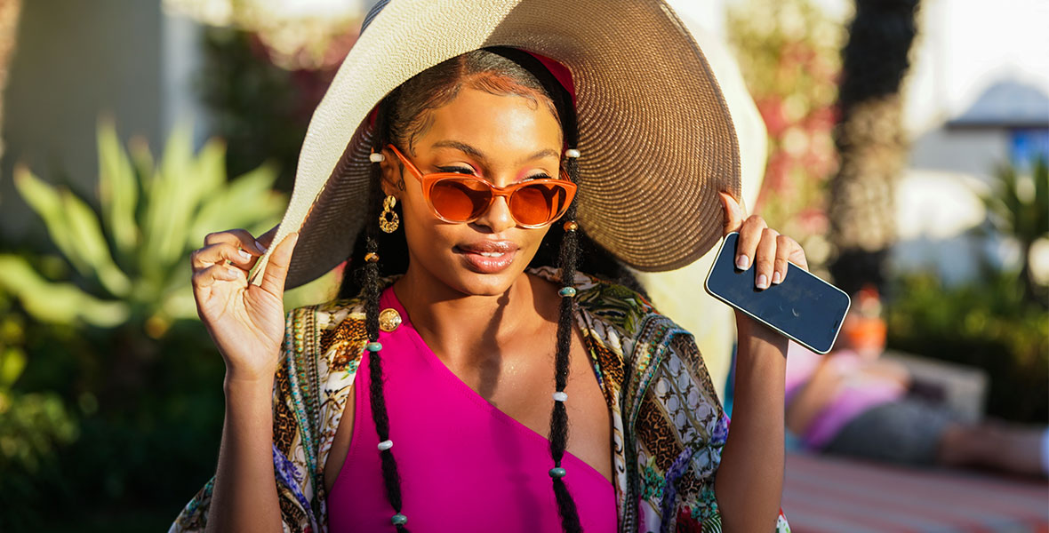 A close-up shot of Zoey Johnson (Yara Shahidi) wearing a large pair of orange sunglasses, hot pink swimsuit, and large sun hat on her head that she’s holding with each hand on grown-ish. Large, green tropical plants are seen in the background.