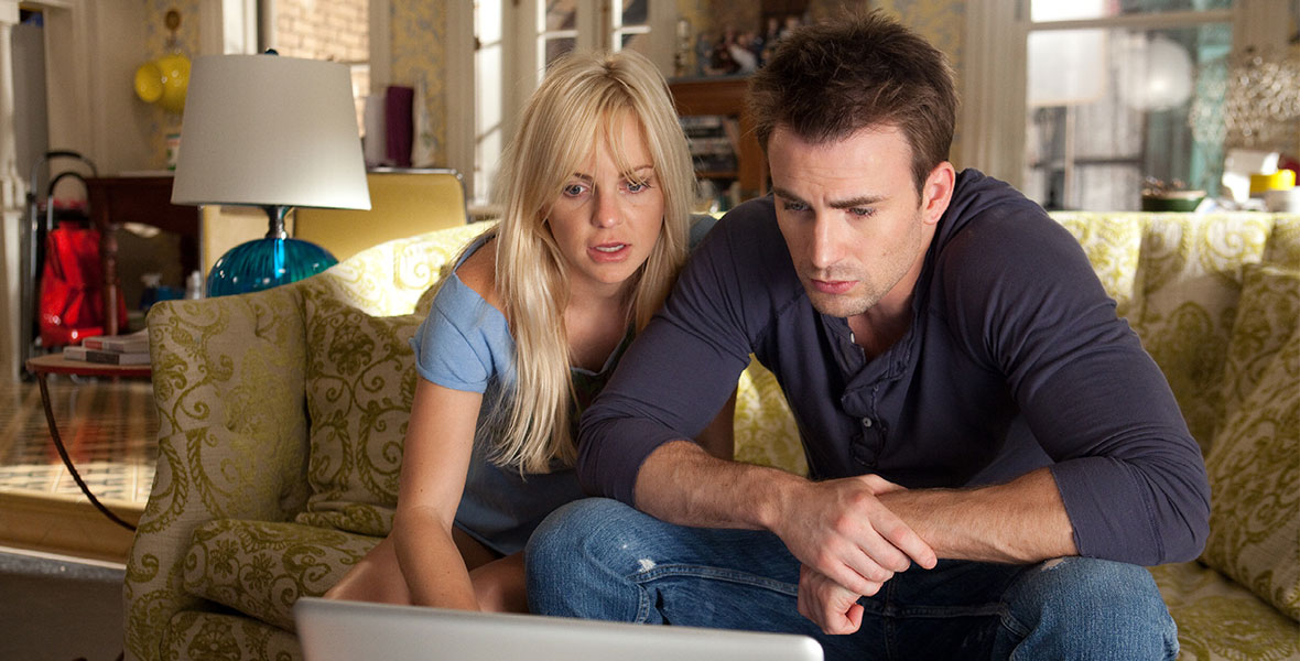 Anna Faris’ Ally Darling, with messy blonde hair in an untidy apartment, sits next to Chris Evans’ Colin Shea, wearing a blue Henley and distressed denim, on a green couch. They are looking at a MacBook screen.