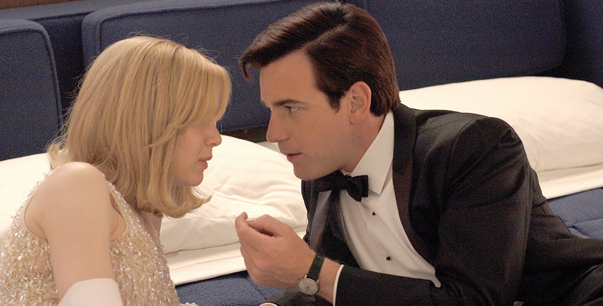 Barbara Novak (Renée Zellweger), dressed in a white beaded gown and gloves, is leaning on a blue upholstered bed next to Catcher Bock (Ewan McGregor), clad in a tux, in an image from Down with Love.