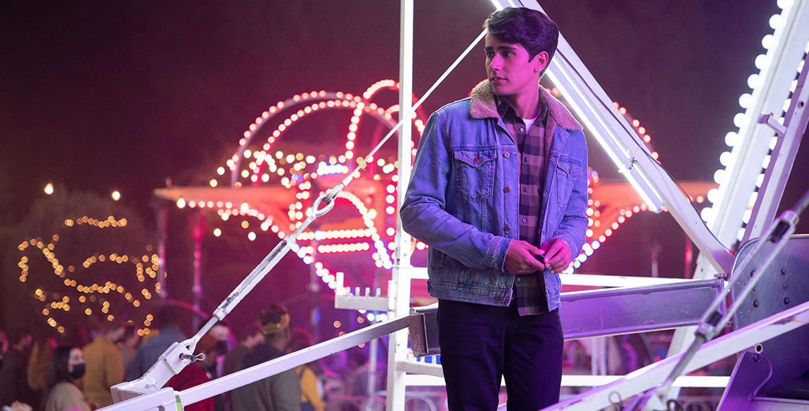 Victor (Michael Cimino) stands on the Ferris Wheel at the winter carnival as he looks on in the series Love, Victor. He’s wearing a plaid shirt, dark slacks, and blue jean jacket.