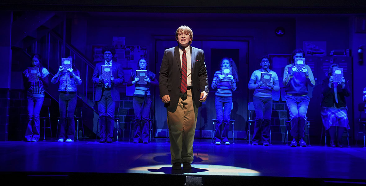 Alt Text: A solo spotlight on Trevor (Holden Hagleberger) in the stage production Trevor: The Musical. He wears a blazer and slacks as he stands center stage in front of a group of fellow performers.