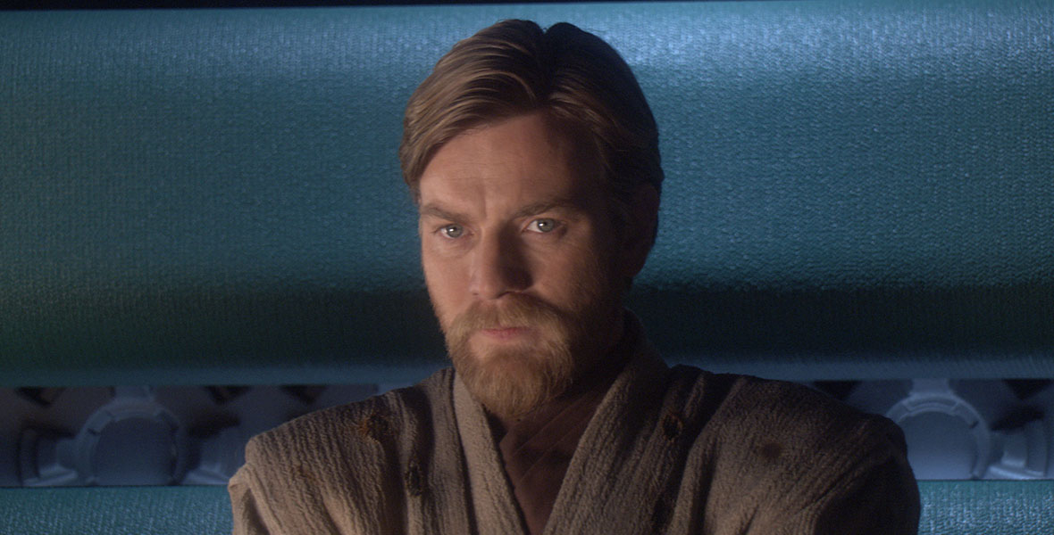 Obi-Wan (Ewan McGregor), in a tan sweater tunic, is looking at something just off camera, set against a blue background, in a still from Star Wars: Revenge of the Sith.