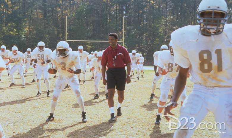 Denzel Washington, playing coach Herman Boone, in Remember the Titans stands smiling in a red polo and black shorts with a whistle around his neck, as football players dressed in white uniforms practice.