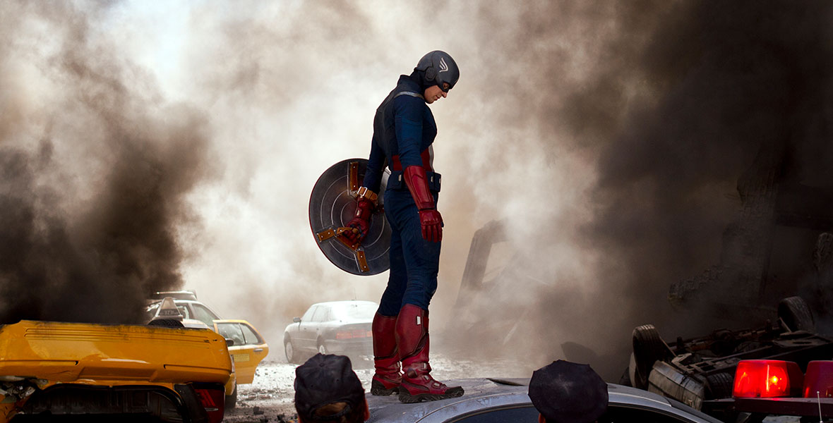 Outfitted as Steve Rogers aka Captain America, wearing red gloves and boots and holding his Vibranium shield, Chris Evans looks to the ground during the Battle of New York in Marvel’s The Avengers. There is smoke in the air and debris on the ground; cars nearby are damaged, and passersby are looking in Captain America’s direction.