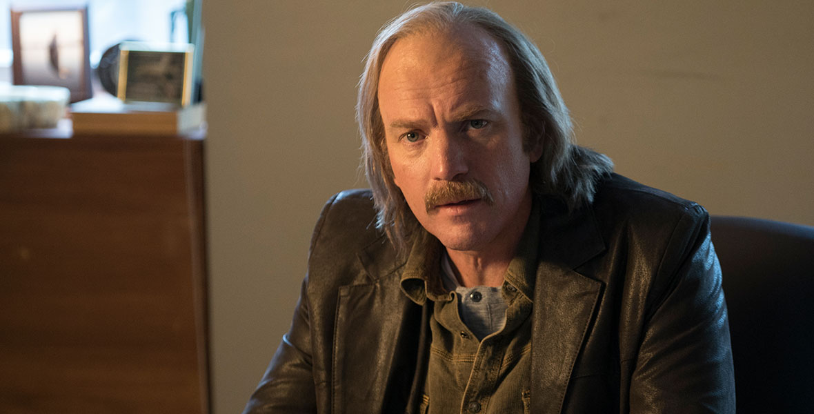 Ray Stussy (Ewan McGregor) has shoulder-length graying hair that’s starting to thin, and he’s wearing a leather jacket with an army green button-up shirt underneath, in an image from season three of FX’s Fargo. He’s sitting at a desk in an office; there’s a coffee cup next to him, and several photo frames on a credenza behind him.