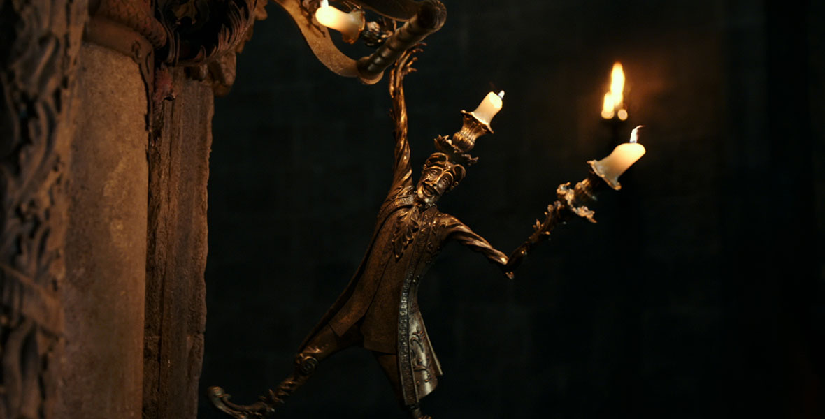 Lumiere (voice of Ewan McGregor) is an anamorphic golden candelabra, and he’s hanging off the stone wall inside the Beast’s castle in a still from Disney’s live-action Beauty and the Beast. There are lit candles coming out of the top of his head and from both of his arms.