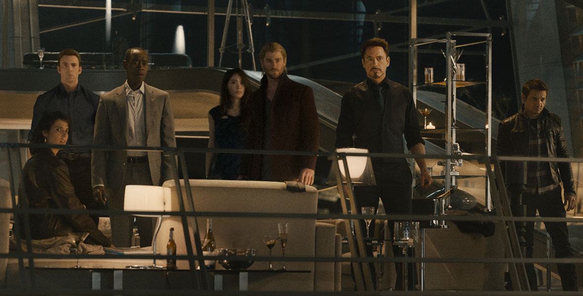 In Avengers: Age of Ultron, the Avengers and their friends stare at Ultron, who is out of frame and has just come alive. Cobie Smulders’ Maria Hill is seated and wears a leather jacket; Chris Evans’ Steve Rogers is standing and wears a blue button-up shirt; Don Cheadle’s James Rhodes is standing with his mouth agape and wears a gray sportscoat; Claudia Kim’s Helen Cho wears a black dress and stands just behind Chris Hemsworth’s Thor, who wears a brown jacket and has his hair pulled back; Robert Downey Jr.’s Tony Stark is standing in the center and wears a dark vest and a dark tie; Jeremy Renner’s Clint Barton stands to his left, wearing a gingham shirt under a black jacket; Mark Ruffalo’s Bruce Banner wears a light blue shirt and a blazer and stands next to Scarlett Johansson’s Natasha Romanoff, who wears a black and white dress.