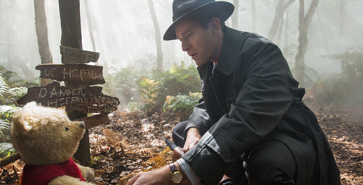 Amidst fog and sun-dappled trees in the Hundred Acre Wood, Winnie the Pooh (voice of Jim Cummings) is wearing his iconic red t-shirt, and is standing in front of several signs labeled “Danger: Heffalumps,” while holding hands with Christopher Robin (Ewan McGregor)—who’s wearing a grey fedora and trench coat and holding a briefcase and umbrella.