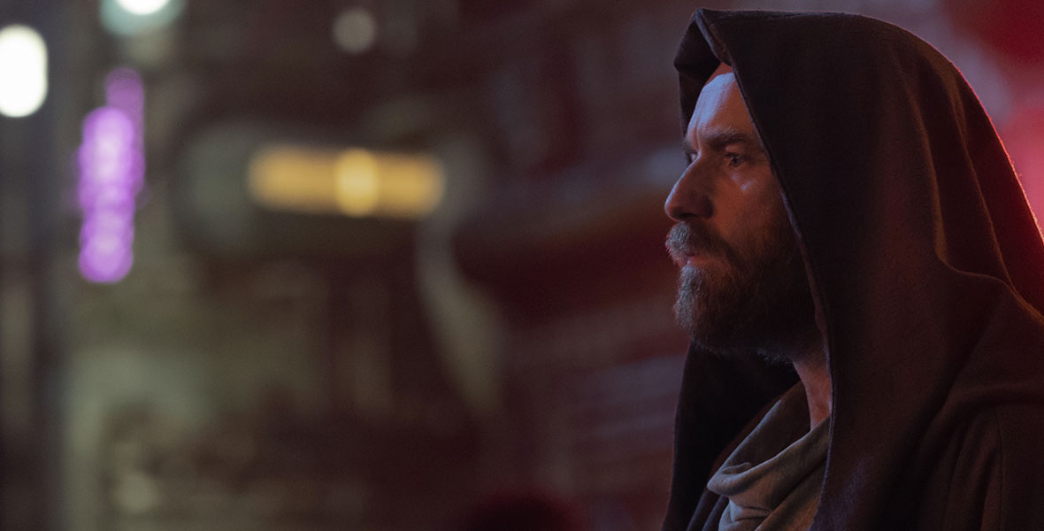 Obi-Wan Kenobi (Ewan McGregor) is wearing a brown cape and cowl and is staring at something off screen, in an image from the Disney+ limited series. There are a few brightly colored, though out-of-focus, lights seen behind him, as well as a few people.