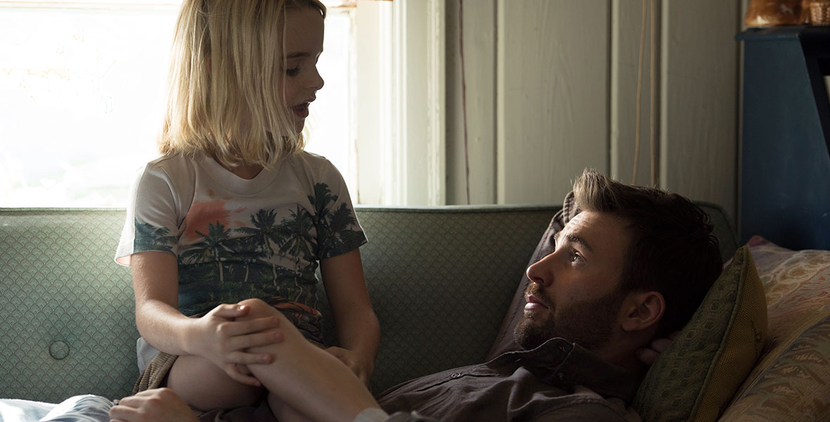 In this scene from Gifted, child prodigy Mary Adler (played by McKenna Grace) is wearing a shirt with palm trees and a sunset on it, paired with khaki corduroys. She has blonde hair and is sitting on the lap of her uncle Frank (played by Chris Evans), who is reclined on the sofa. Frank is wearing a brown shirt and is listening intently to his niece.