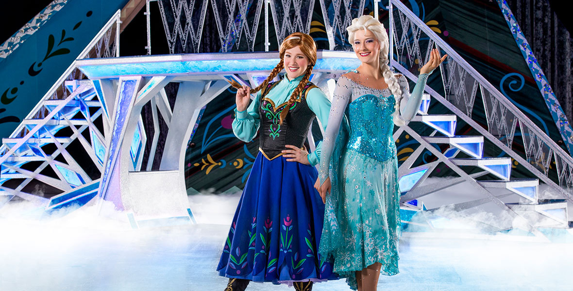 Anna and Elsa stand back to back on a Disney On Ice stage. They are wearing ice skates and have a large staircase made of ice behind them.